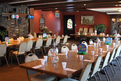 A dining room with multiple rectangular tables arranged in rows, each table set with white cups and napkins. The room has beige cushioned chairs, a brick fireplace on the left wall, and a stained glass door in the background. The walls are painted red and decorated with a mirror, a painting, and a large green plant. Some tables have colorful birthday decorations.
