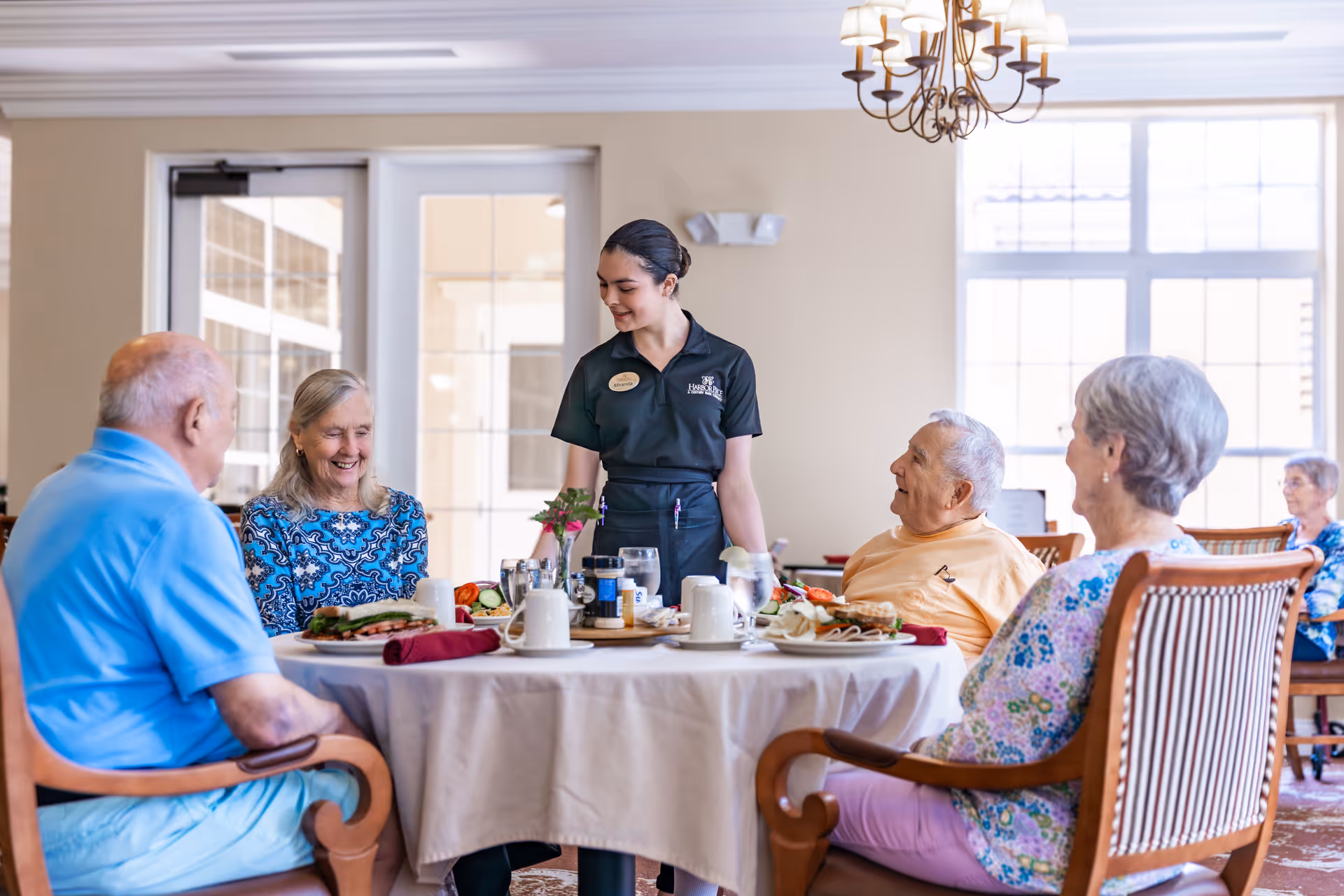 A server attends to four elderly residents seated around a round dining table in a bright dining room.