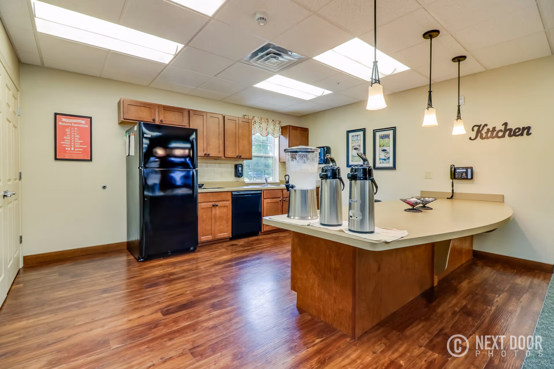 Well-lit communal kitchen with a center island, coffee dispensers, black appliances, and wooden cabinets.