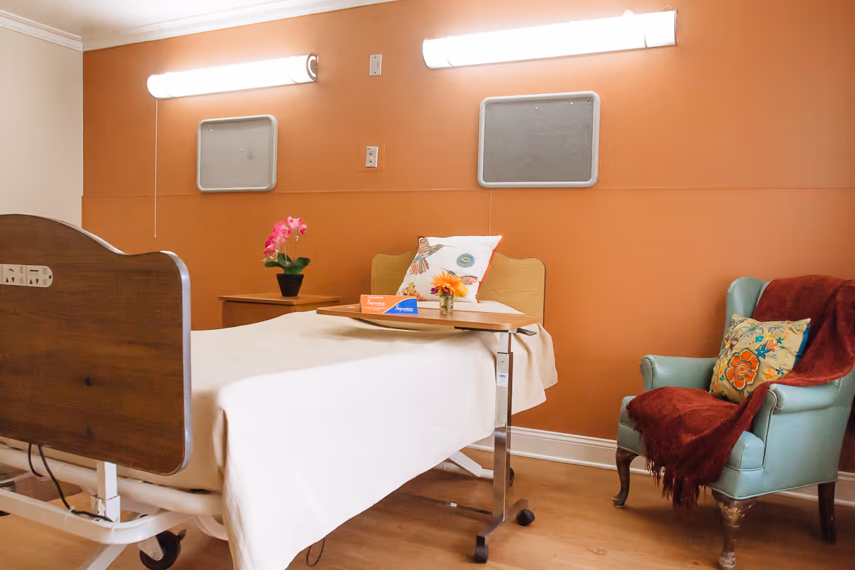 A senior living facility room with a hospital-style bed covered in a white blanket, a small wooden side table with a pink orchid plant, and a light blue armchair with a colorful floral pillow and a red throw blanket. The walls are painted orange with two rectangular panels and two bright wall lights above the bed.