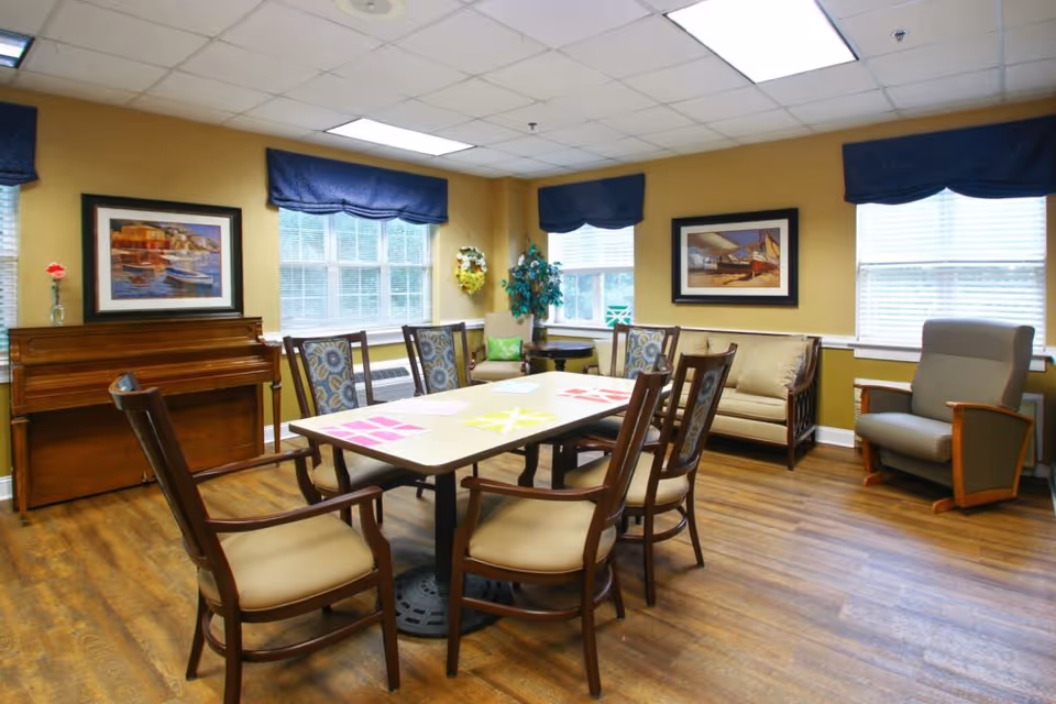 Bright senior living common room with a central table and chairs, a piano, sofa and armchair beneath windows.