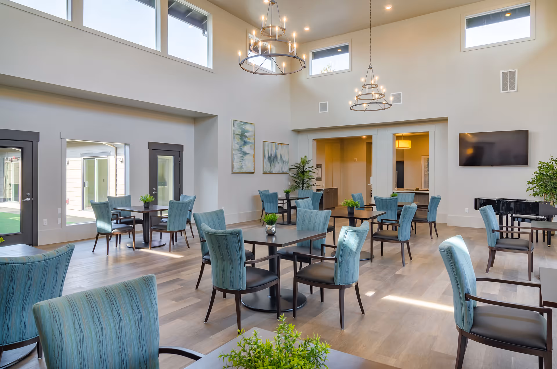 Bright high-ceilinged communal dining/lounge area with multiple tables and teal upholstered chairs, chandeliers, a wall-mounted TV and a piano.