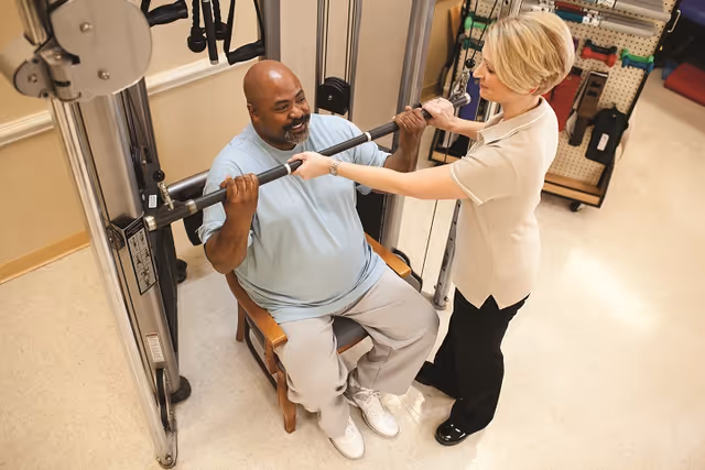 A man seated on a wooden chair is using a lat pulldown machine for exercise while a woman stands beside him, assisting and supporting the exercise in a rehabilitation or fitness room.