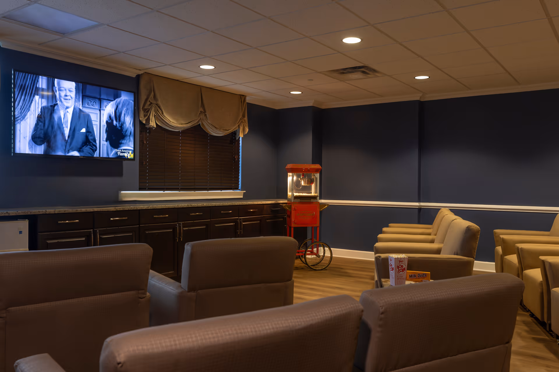 A cozy entertainment room with multiple beige armchairs arranged facing a wall-mounted television playing a black-and-white show. The room has dark blue walls with white trim, a window with closed blinds and a beige valance, dark wood cabinets with a granite countertop, and a vintage-style red popcorn machine on wheels in the corner.