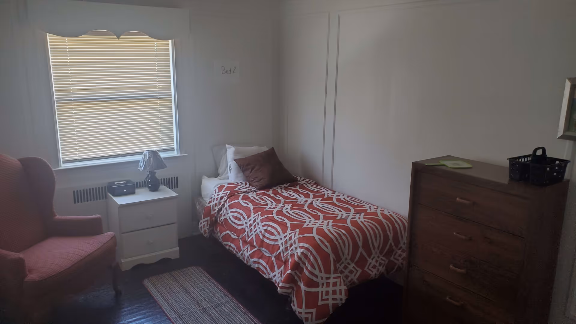 A small bedroom with a single bed covered in a red and white patterned comforter. There is a brown pillow and a white pillow on the bed. Next to the bed is a white nightstand with a lamp and a small black device on it. A window with closed blinds is above the nightstand. To the left of the nightstand is a red upholstered armchair. On the right side of the room is a wooden dresser with four drawers and a black basket on top. The walls are plain white, and there is a small sign on the wall above the bed that reads 'Bed 2'.