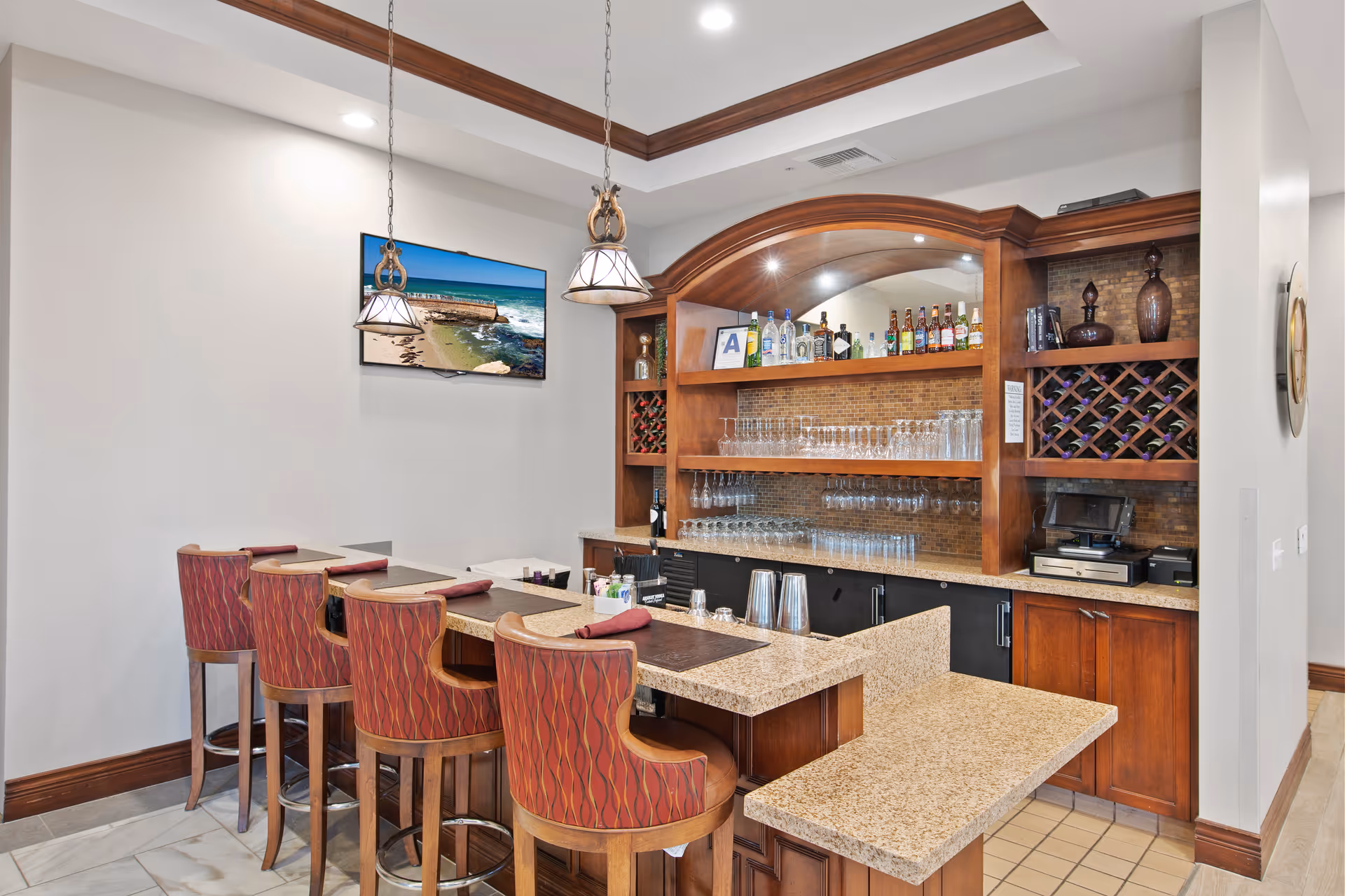 Indoor bar area with a granite countertop, upholstered bar stools, wood cabinetry and shelves stocked with glassware and bottles.
