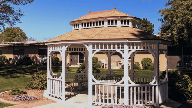 A white wooden gazebo on a sunny grassy courtyard with a building and shrubs in the background.