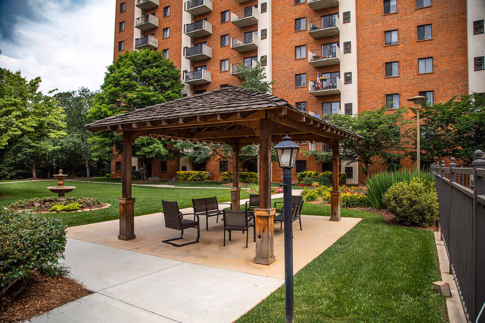 A wooden gazebo with patio chairs sits on a concrete pad in a landscaped courtyard in front of a multi-story brick residential building.