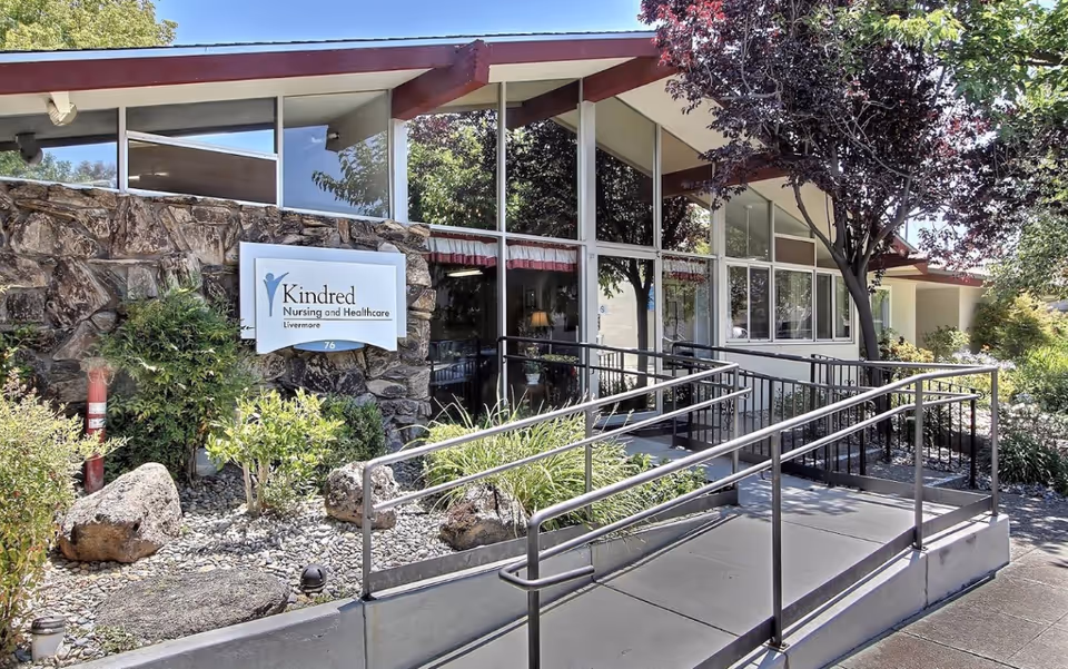 Exterior view of a healthcare facility with a stone wall and a sign that reads 'Kindred Nursing and Healthcare Livermore'. The building has large windows, a ramp with metal railings, and surrounding greenery including trees and bushes.