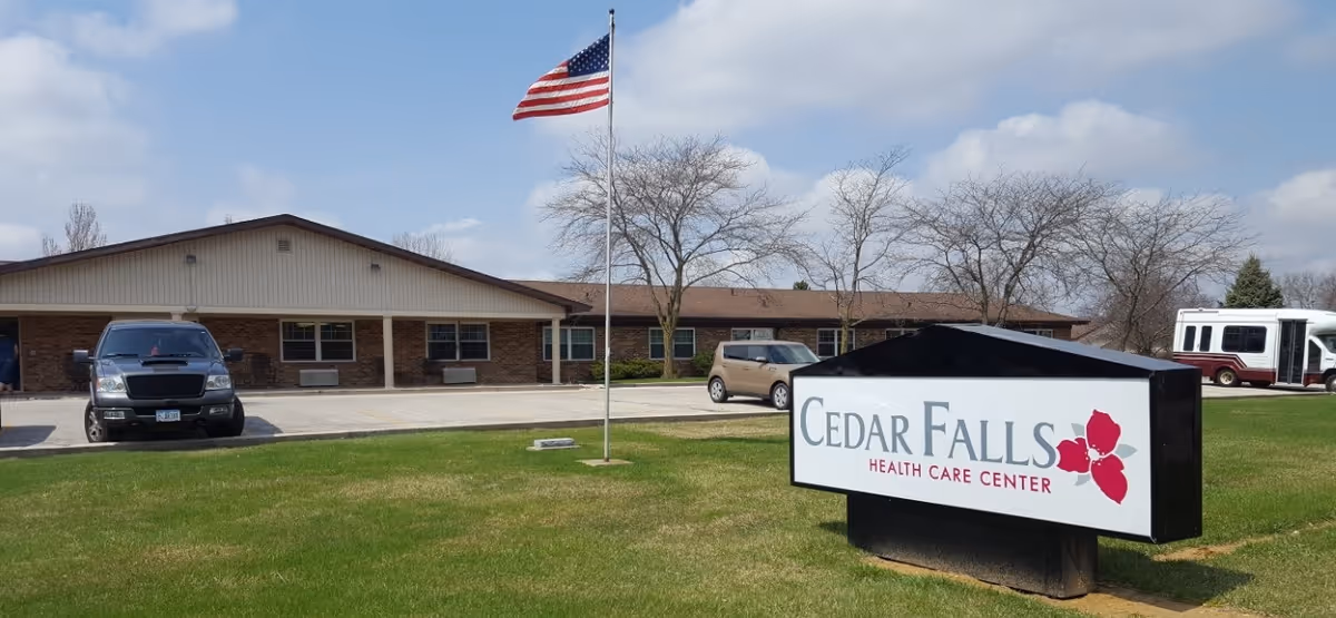 Exterior view of Cedar Falls Health Care Center building with a parking lot, several vehicles, an American flag on a flagpole, and a sign displaying the facility's name on a grassy area.