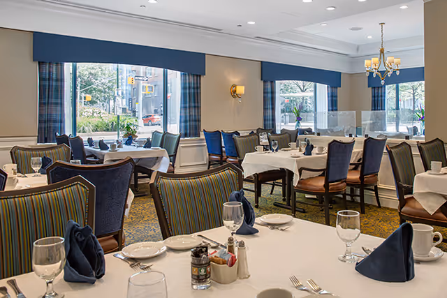 A bright and elegant dining room with multiple tables covered in white tablecloths, set with plates, glasses, silverware, and navy blue folded napkins. The room features large windows with blue valances and plaid curtains, allowing natural light to fill the space. Comfortable chairs with striped and solid upholstery surround the tables, and a chandelier and wall sconces provide additional lighting.