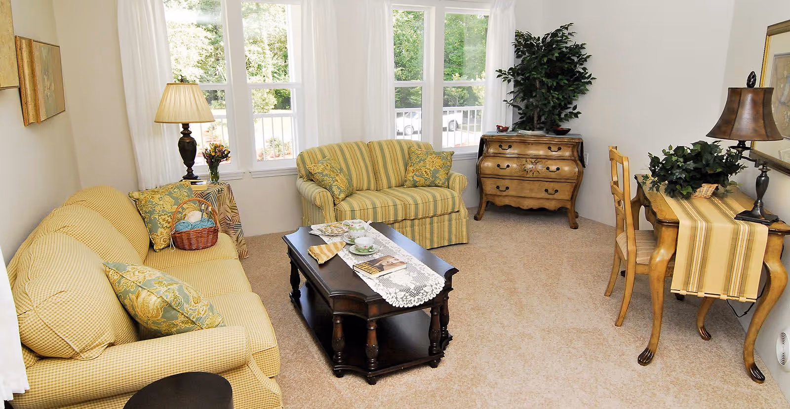 A cozy living room with two yellow patterned sofas, a dark wooden coffee table with a lace runner, teacups, and a book. There is a wooden side table with a lamp and flowers, a wooden dresser with a plant on top, and a wooden desk with a chair, lamp, and greenery. Large windows with white curtains allow natural light to fill the room.