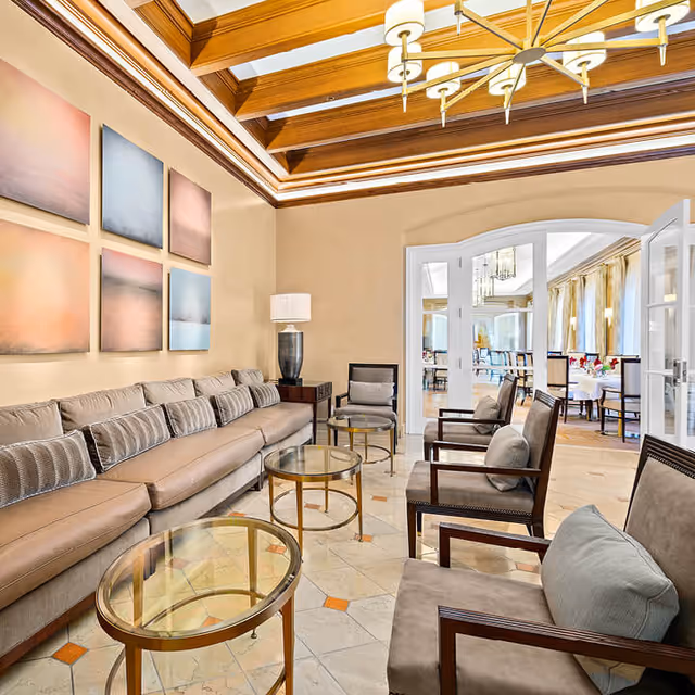 A bright and elegant living room area with a long beige sofa adorned with multiple cushions, three armchairs with light-colored cushions, and three glass-top round coffee tables with gold frames. The room features a coffered wooden ceiling with a modern chandelier, beige walls with six abstract paintings, and a table lamp on a side table. In the background, through open double doors, a dining area with tables and chairs is visible.