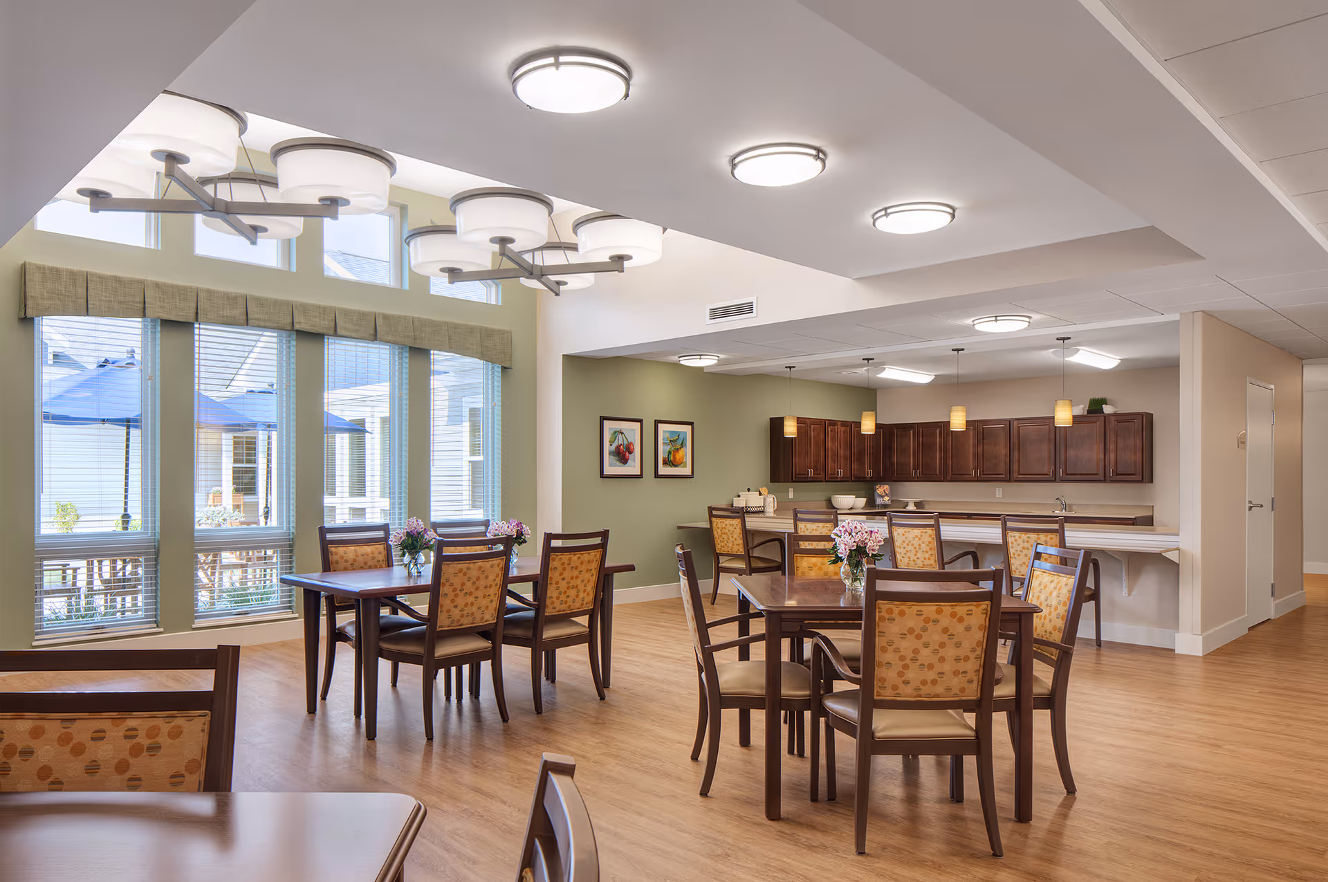 Bright communal dining room with several tables and chairs, large windows, and an open kitchen area.