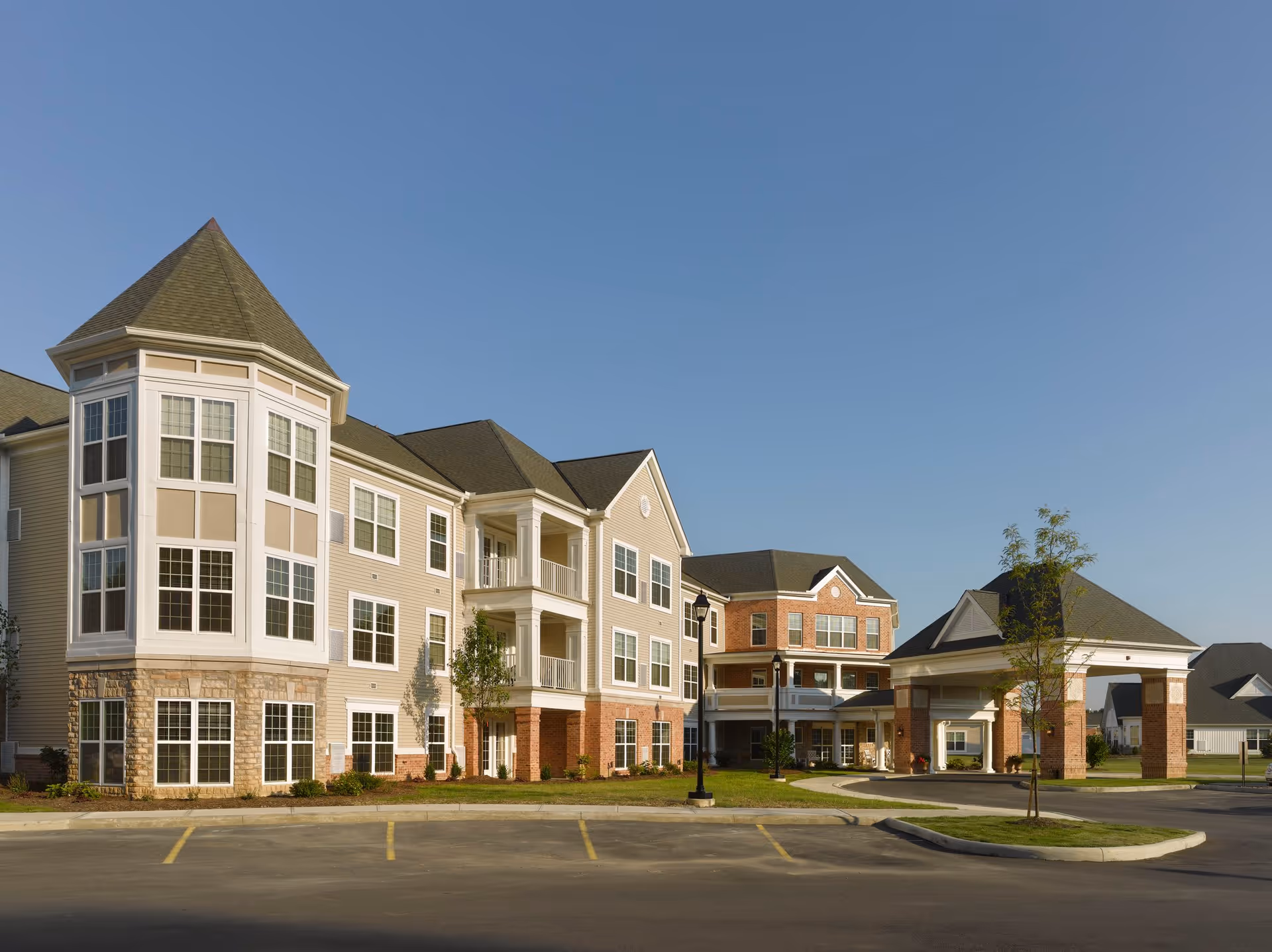 Exterior view of a multi-story senior living facility building with beige and brick facade, large windows, and a covered entrance driveway under a clear blue sky.
