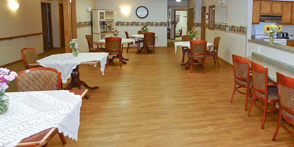 A spacious dining area with wooden floors and several tables covered with white lace tablecloths. Each table has a vase with flowers. There are wooden chairs around the tables and a counter with additional chairs on the right side. The room has soft lighting, a clock on the wall, and a kitchen area visible in the background.