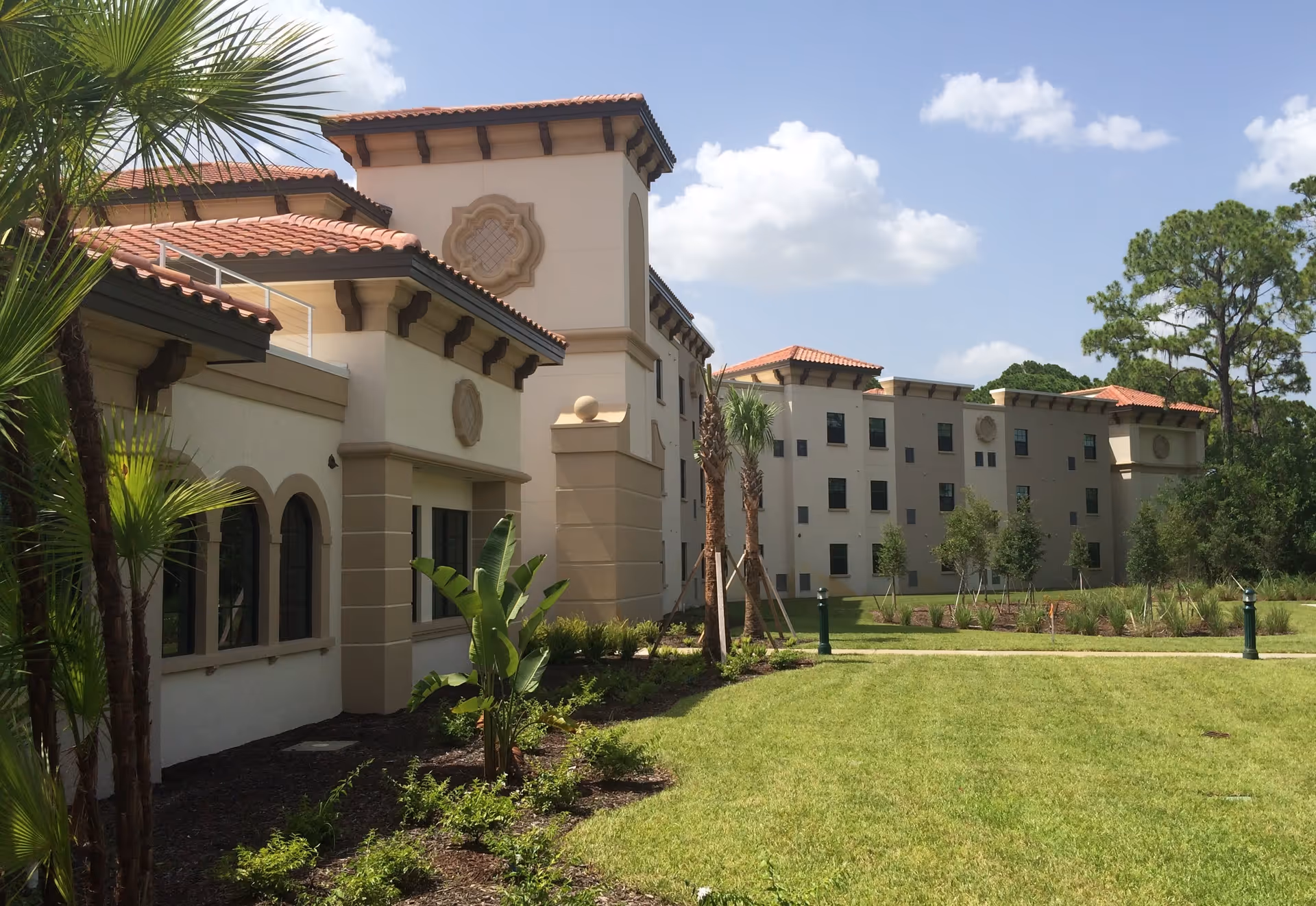 Exterior view of a multi-story senior living facility building with beige walls and red-tiled roofs, surrounded by green grass, palm trees, and other landscaping under a partly cloudy sky.
