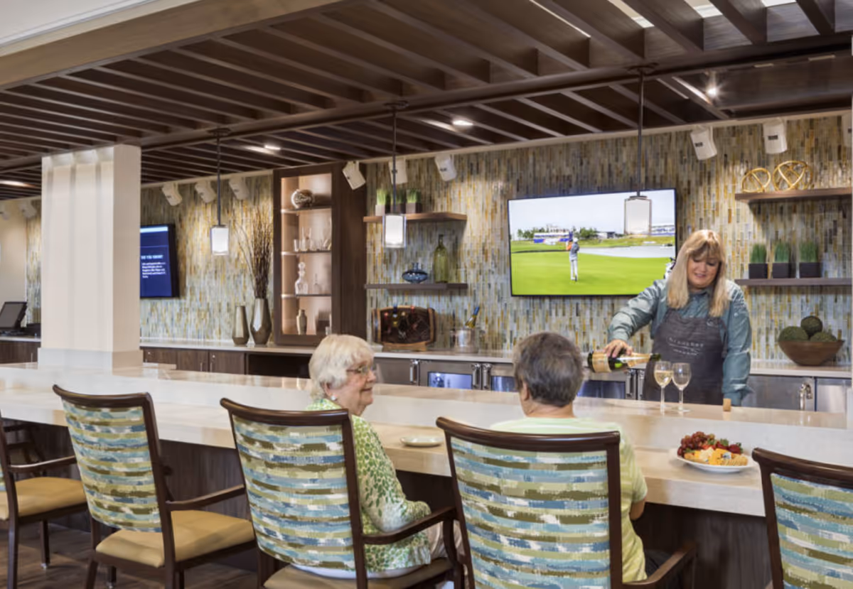 Residents sit at a bar counter while a staff member pours wine in a decorated senior living common dining area.