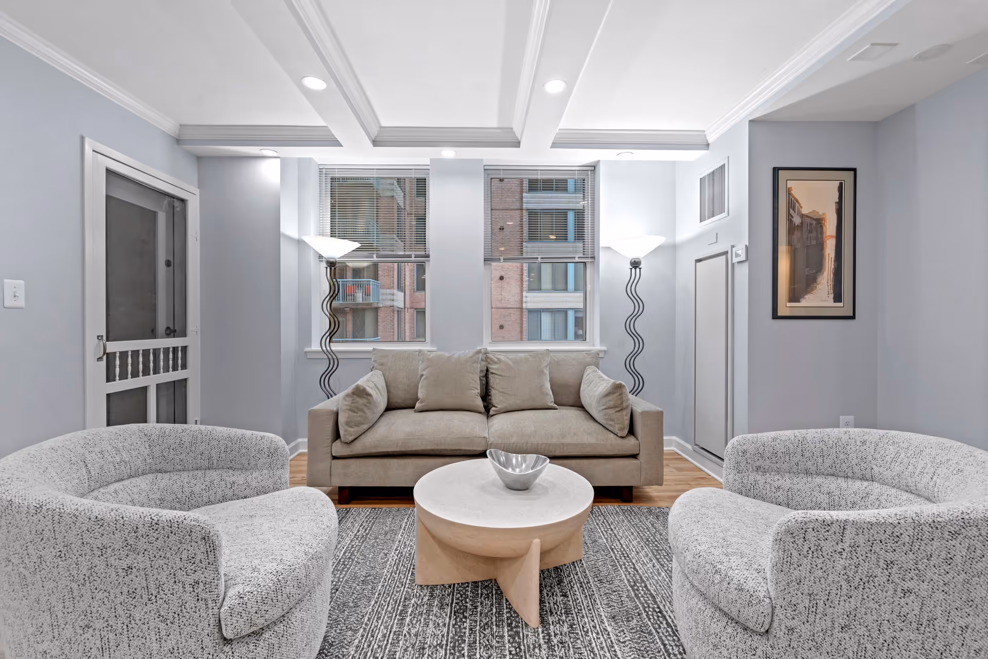 A modern living room with a beige sofa and two curved gray armchairs arranged around a round white coffee table with a decorative bowl. The room features light gray walls, two tall windows with blinds, two floor lamps with wavy stands, a patterned rug, and a framed artwork on the right wall.