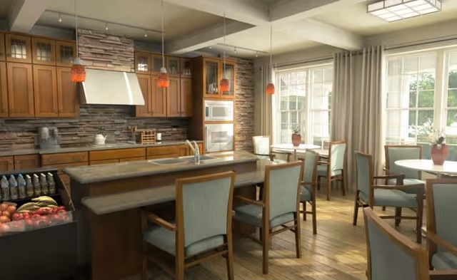 A spacious dining area with wooden floors and large windows letting in natural light. The room features multiple round tables with chairs, a kitchen island with seating, wooden cabinetry, and modern pendant lights hanging from the ceiling. There is a stone backsplash behind the kitchen area and fresh fruit displayed on the counter.