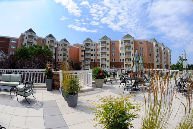 Outdoor patio area with tables, chairs, umbrellas, and potted plants in front of a multi-story residential building under a partly cloudy sky.