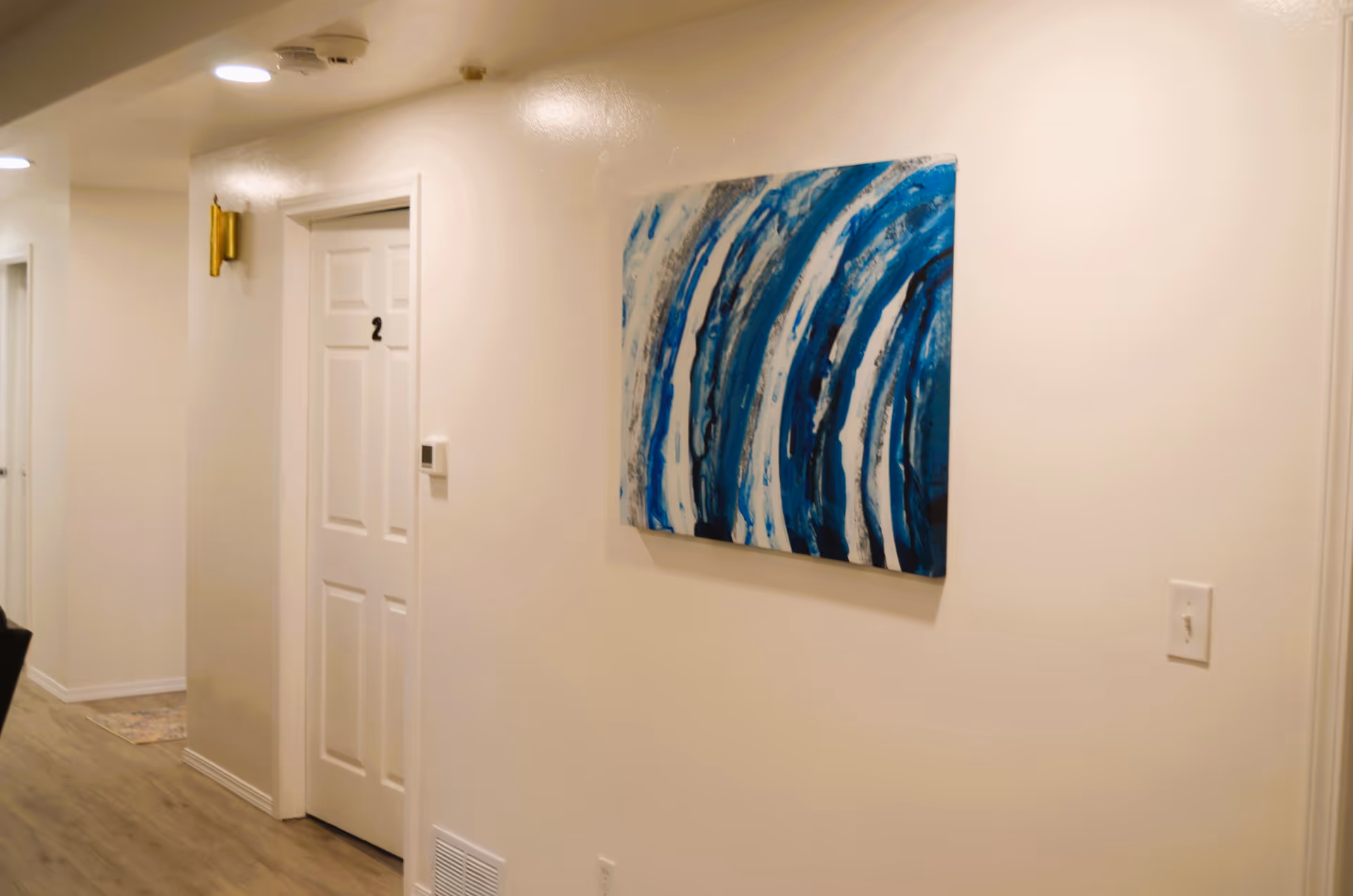 Interior hallway of a senior living facility with white walls and wooden flooring. There are white doors numbered 2 and another door further down the hall. A blue and white abstract painting is hung on the wall, and ceiling lights illuminate the corridor.