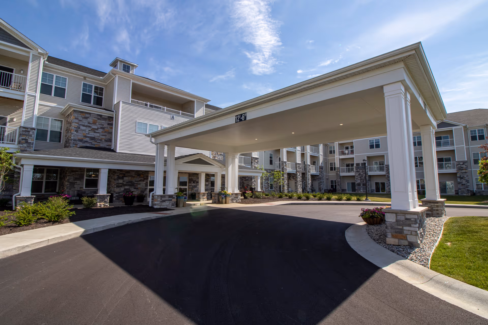 Covered porte-cochère entrance of a multi-story senior living building with stone and siding facade under a blue sky.