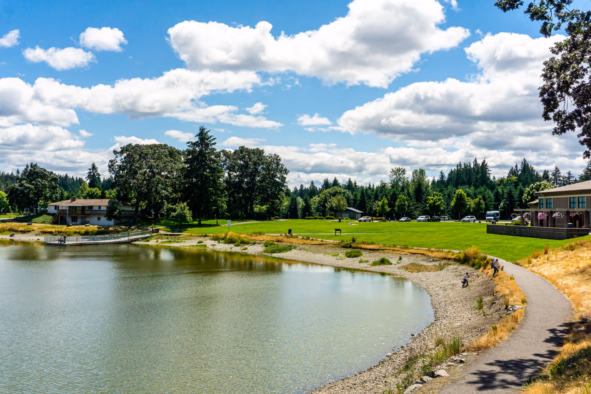 Lakeside view with a walking path, grassy lawn, trees and buildings under a blue sky with clouds.