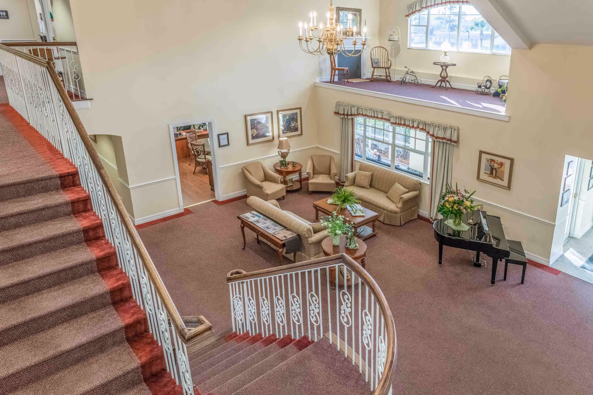 Spacious two-story interior lounge with a curved carpeted staircase, seating area with sofas and chairs, a grand piano, and a chandelier overlooking a mezzanine.