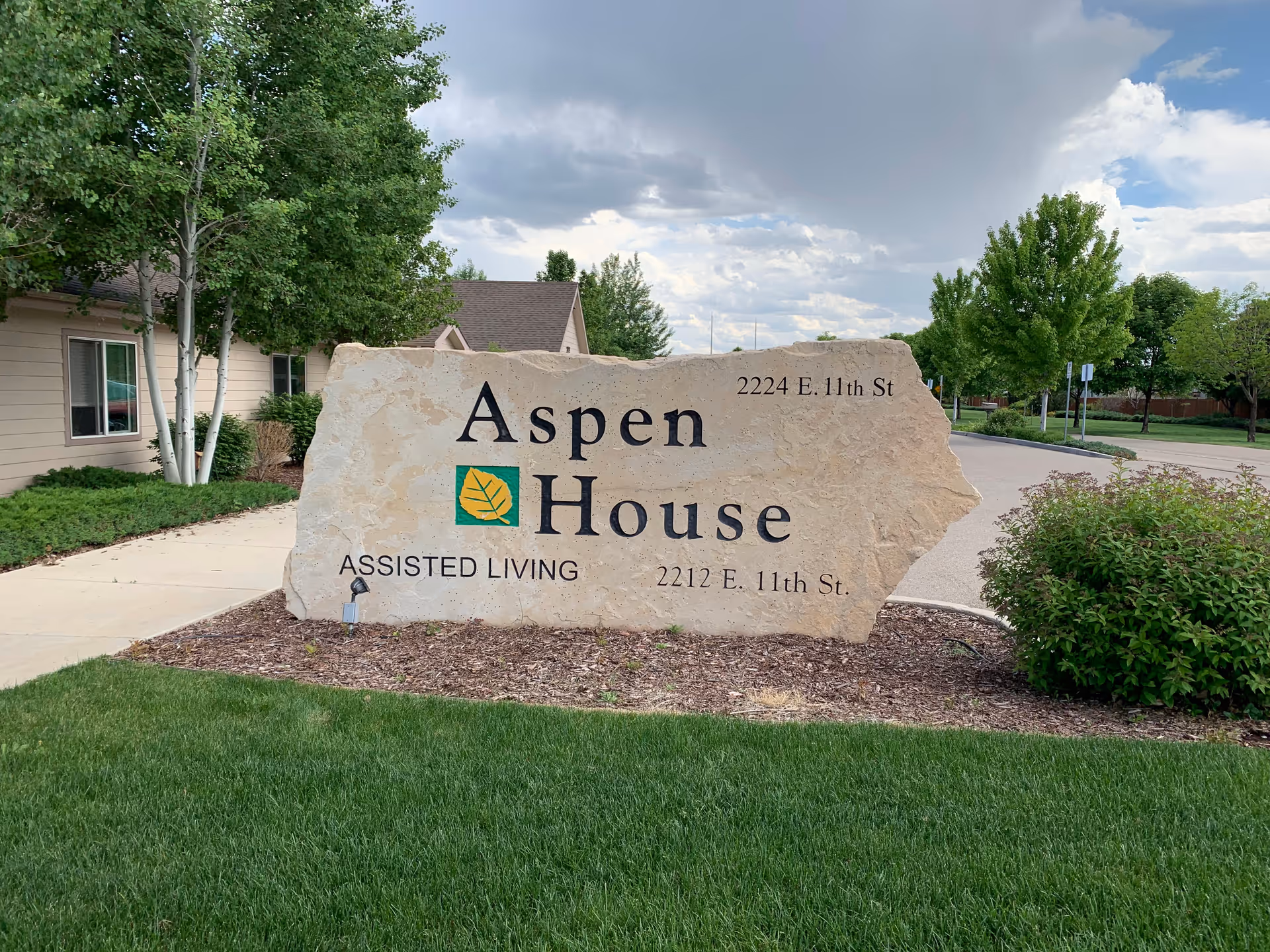 Large stone sign for Aspen House Assisted Living with addresses 2224 E. 11th St and 2212 E. 11th St, surrounded by green grass, bushes, trees, and a sidewalk under a partly cloudy sky.