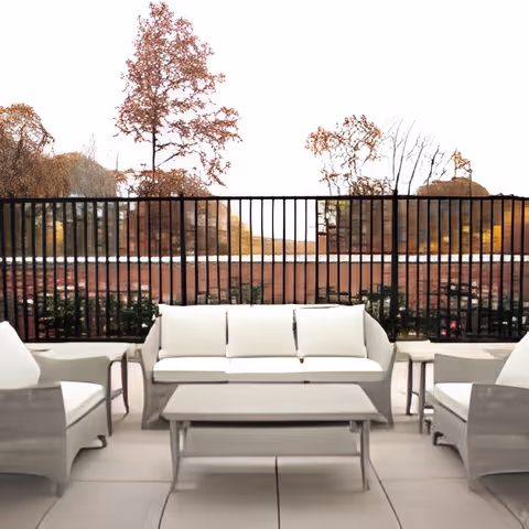 Outdoor patio with white cushioned seating and a coffee table facing a black metal railing and trees beyond.