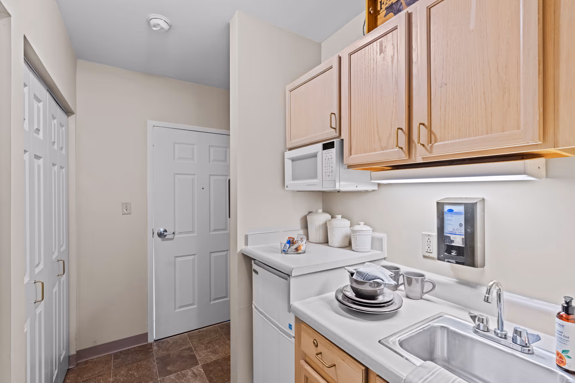 Compact kitchenette with a sink, microwave, wooden cabinets, countertop dishes and a closed door at the end of a short hallway.