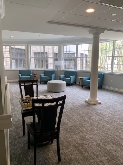 A bright and spacious sitting area with large windows letting in natural light. The room features four blue armchairs arranged in a semi-circle around a round white ottoman. There are two small side tables with lamps and decorative items. In the foreground, there are two black chairs facing a small table with a chessboard. The room has light gray carpet and a white column near the center.