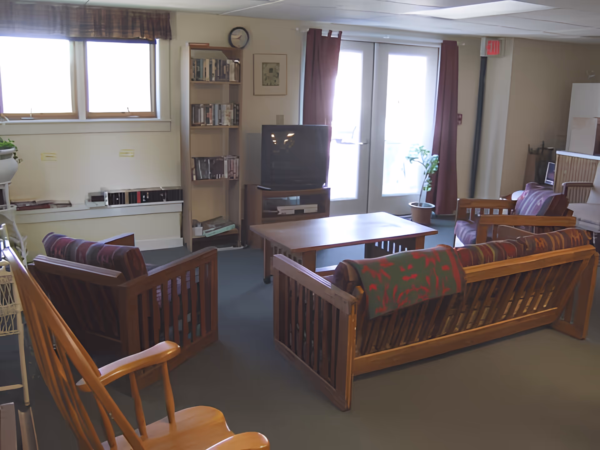 A cozy common living room with wooden sofas and chairs arranged around a coffee table, a TV and bookshelf, and double doors to the outside.