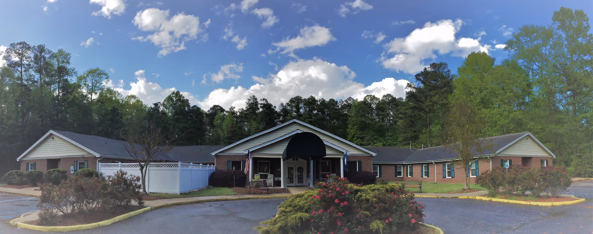 Exterior view of Dominion Village at Williamsburg, a single-story brick building with a central entrance covered by a black awning. The building is surrounded by trees and bushes, with a circular driveway and parking area in front under a partly cloudy blue sky.