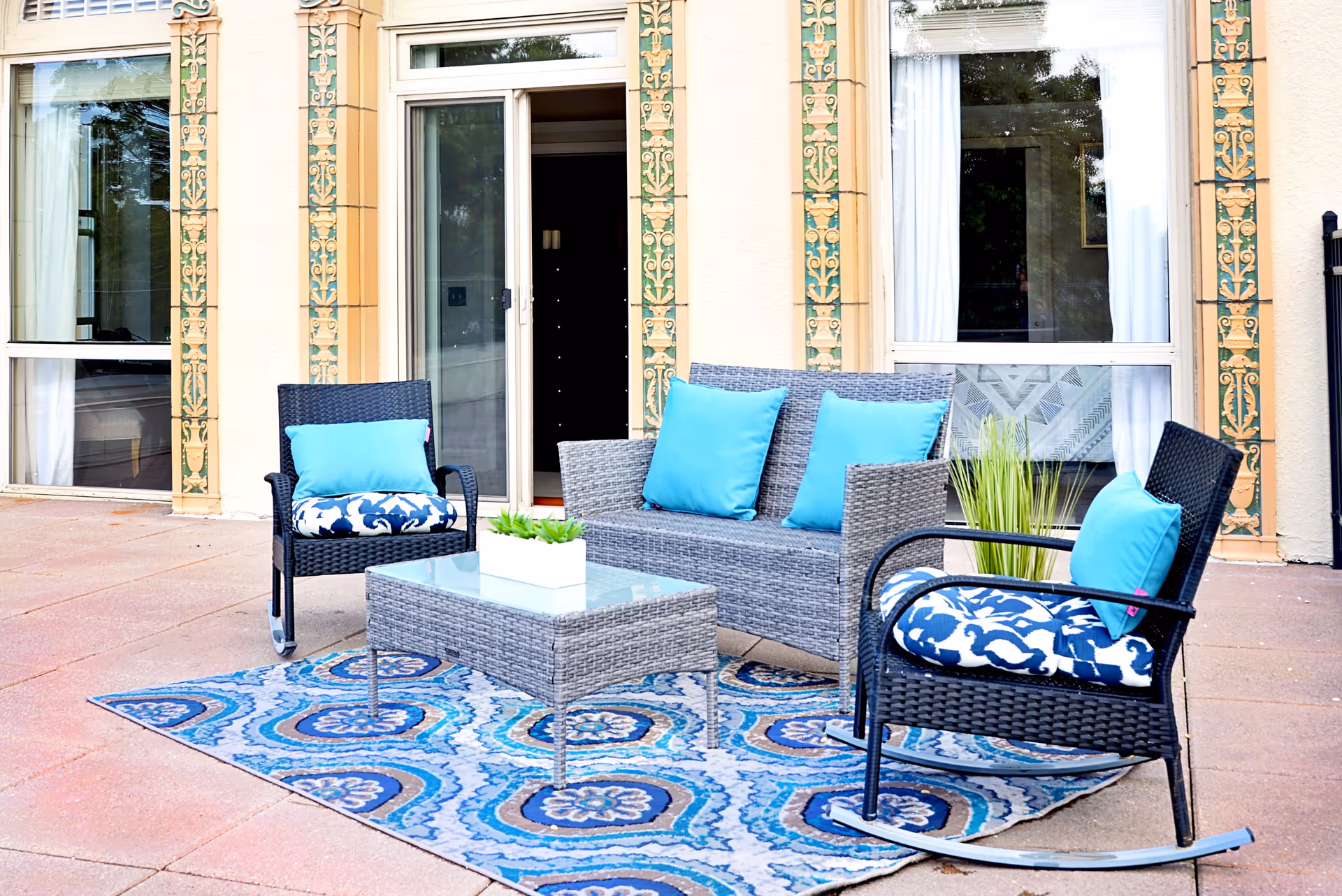 Outdoor patio area with a gray wicker loveseat and two matching rocking chairs, all with blue and white cushions. A glass-top coffee table sits on a blue patterned outdoor rug. The patio is adjacent to a building with large windows and decorative trim around the door and windows.