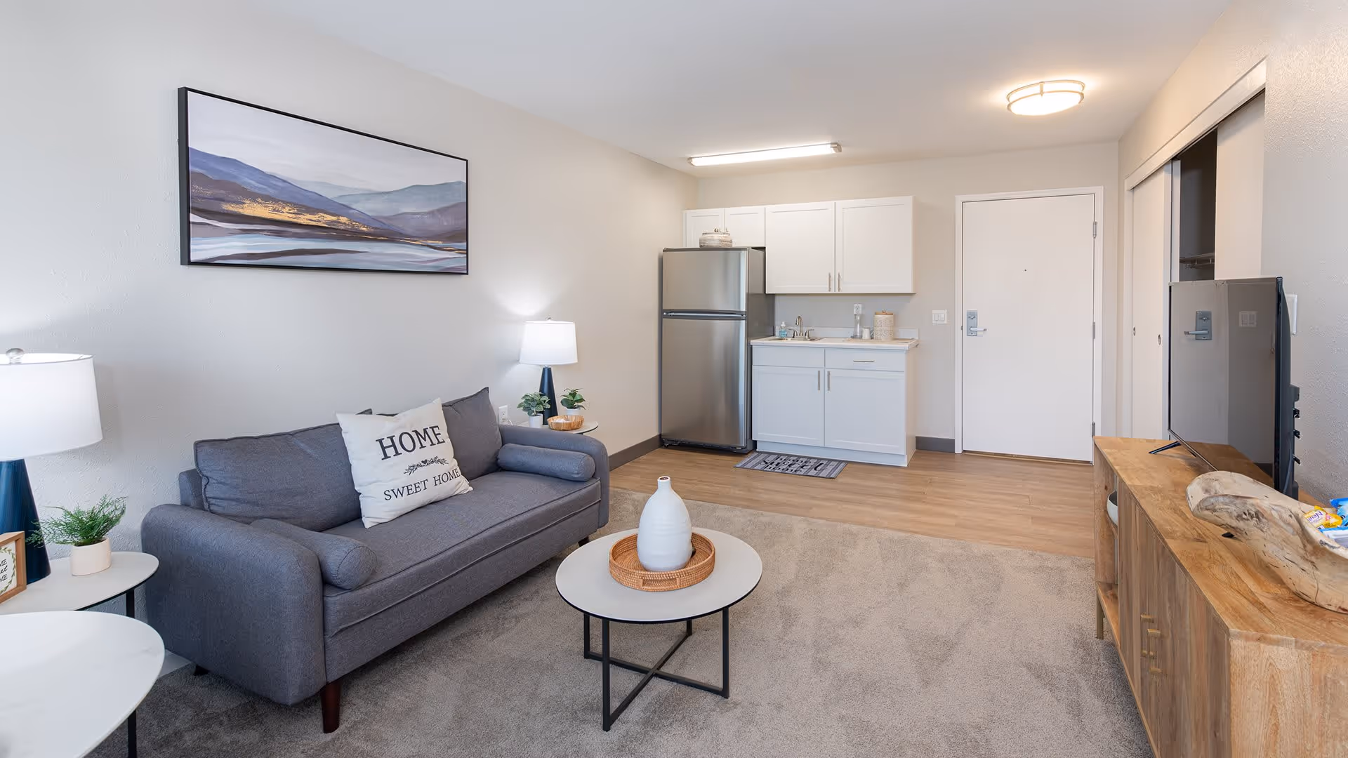 A cozy living area with a gray sofa adorned with a 'Home Sweet Home' pillow, a round coffee table with a decorative vase, and a wooden TV stand with a flat-screen TV. The room features light-colored walls, a framed landscape painting above the sofa, and a small kitchenette with white cabinets, a stainless steel refrigerator, and a sink. The floor transitions from carpet in the living area to wood in the kitchenette. There are two table lamps providing warm lighting.