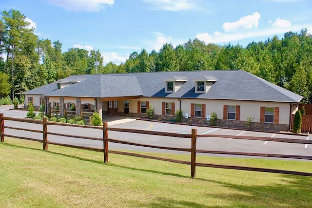 Front exterior of a single-story assisted living building with a covered entrance, dormer windows, parking area, and a wooden fence with trees in the background.