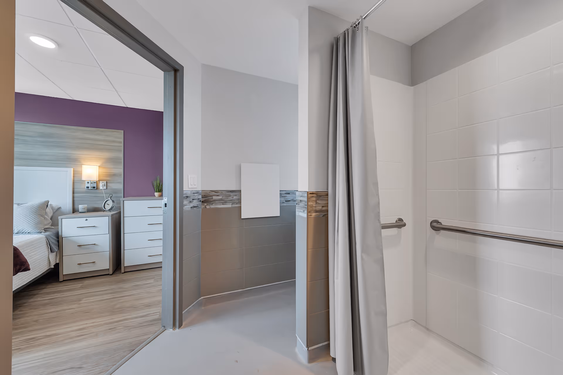 Accessible bathroom with a tiled roll-in shower and grab bars visible next to a bedroom with nightstands.