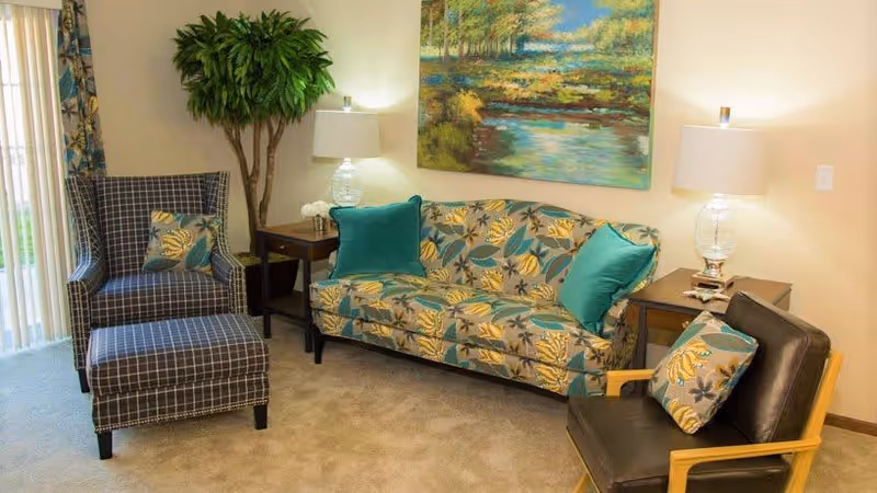 A cozy living room area featuring a patterned sofa with teal and yellow floral cushions, a matching armchair with an ottoman, a dark leather chair with a floral cushion, two side tables with glass lamps, a large green potted plant, and a landscape painting on the wall.