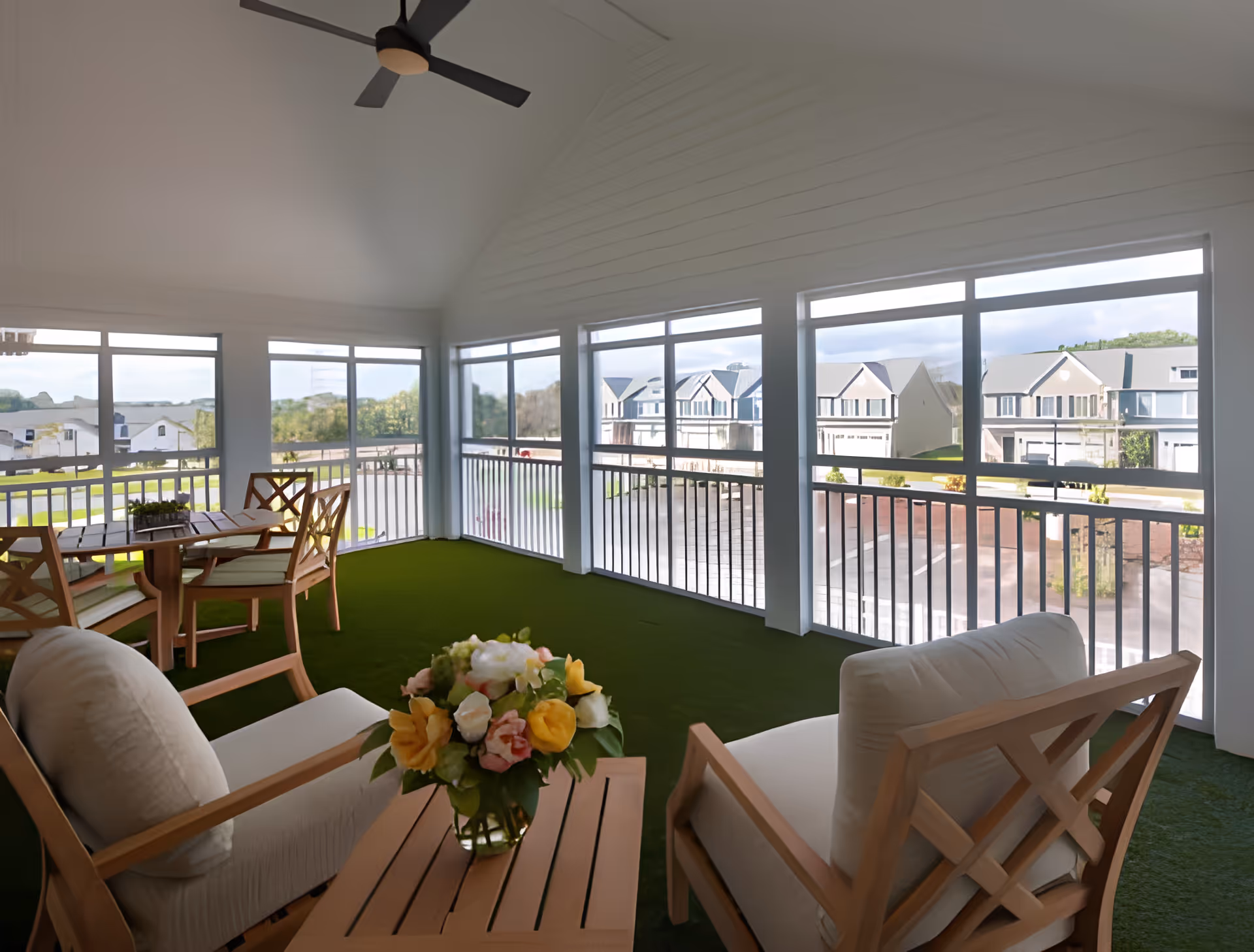A furnished screened-in sunroom/porch with cushioned chairs, a dining table, ceiling fan, and views of neighboring houses.