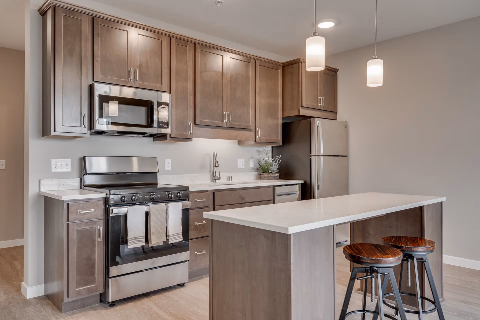 Modern kitchen with wooden cabinets, stainless steel appliances including a microwave, stove, and refrigerator, a white countertop island with two wooden stools, and pendant lights hanging from the ceiling.