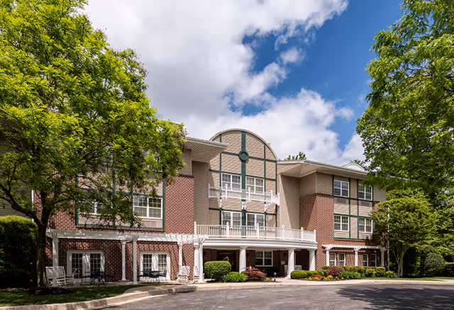 Exterior view of a three-story senior living facility building with a mix of brick and siding facade, white railings on balconies, surrounded by green trees and shrubs under a partly cloudy blue sky.