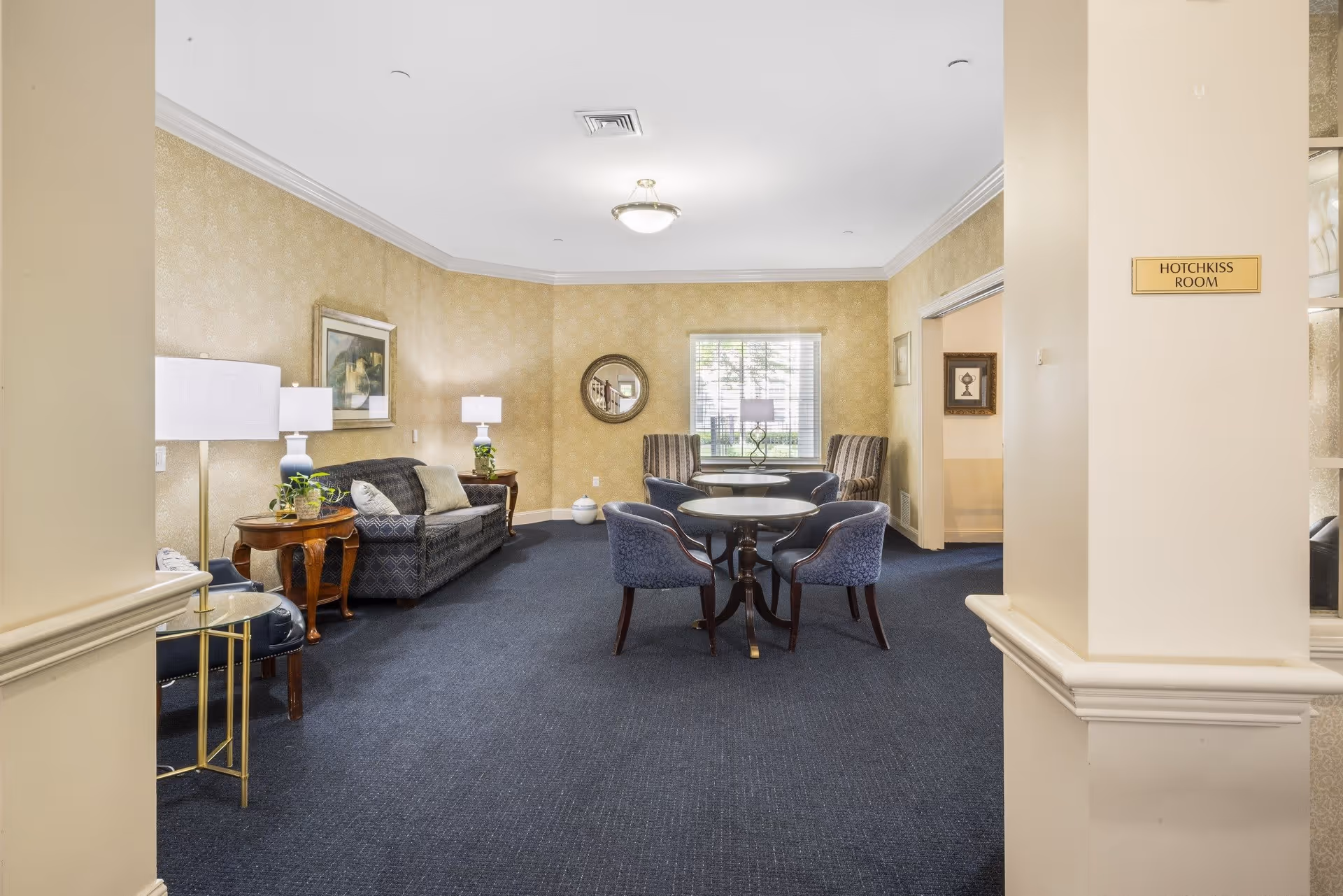 A cozy sitting room named Hotchkiss Room with a dark blue carpet, beige patterned wallpaper, and a ceiling light fixture. The room features a dark patterned sofa with cushions, two wooden side tables with lamps and plants, a round table with four blue upholstered chairs, and two striped armchairs near a window with blinds. There are framed pictures and a round mirror on the walls.