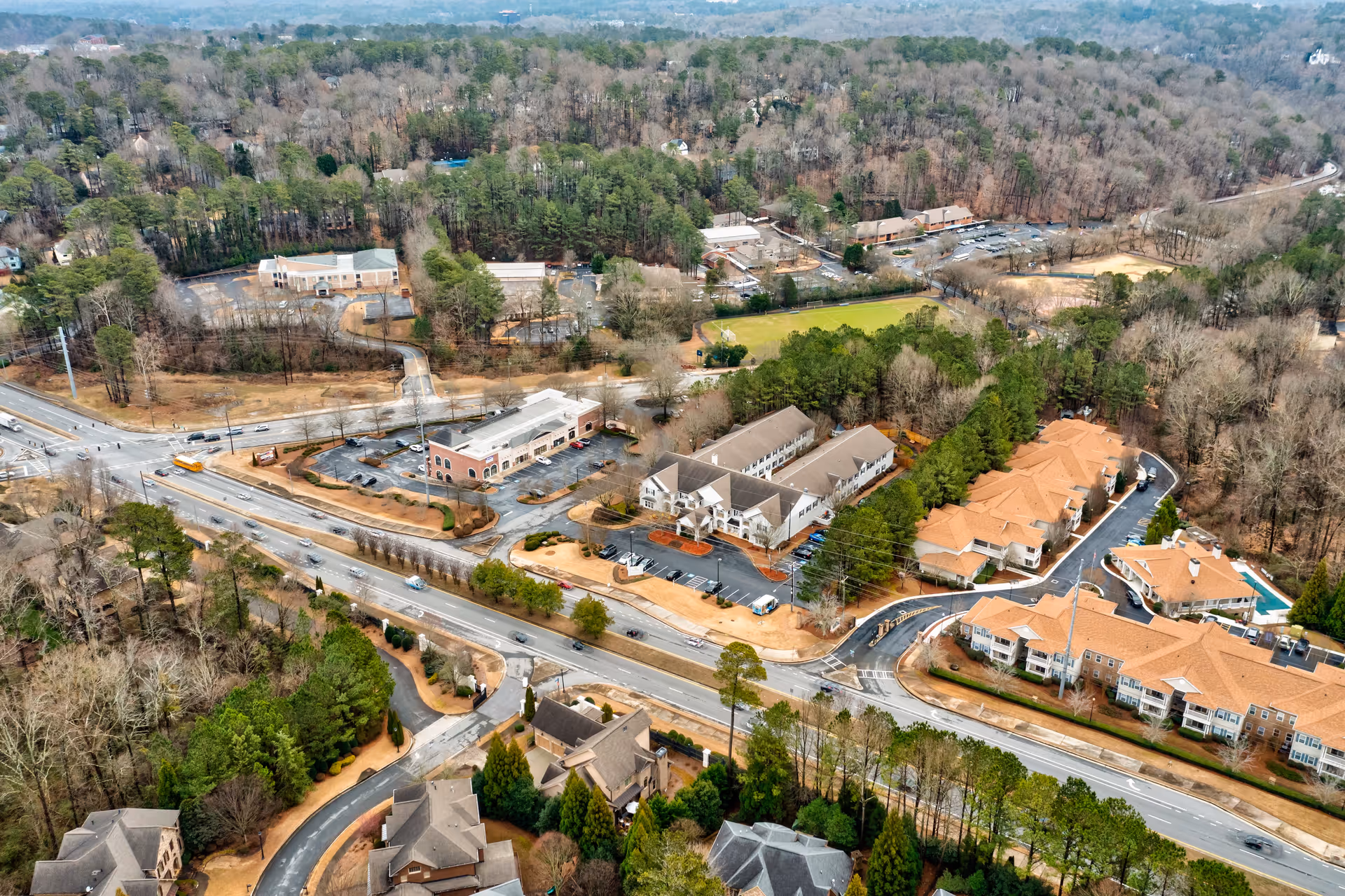 Aerial view of a suburban area showing multiple buildings, roads, parking lots, and surrounding trees. The image includes a mix of residential and commercial buildings with a main road running through the center. The landscape is dotted with patches of greenery and wooded areas.