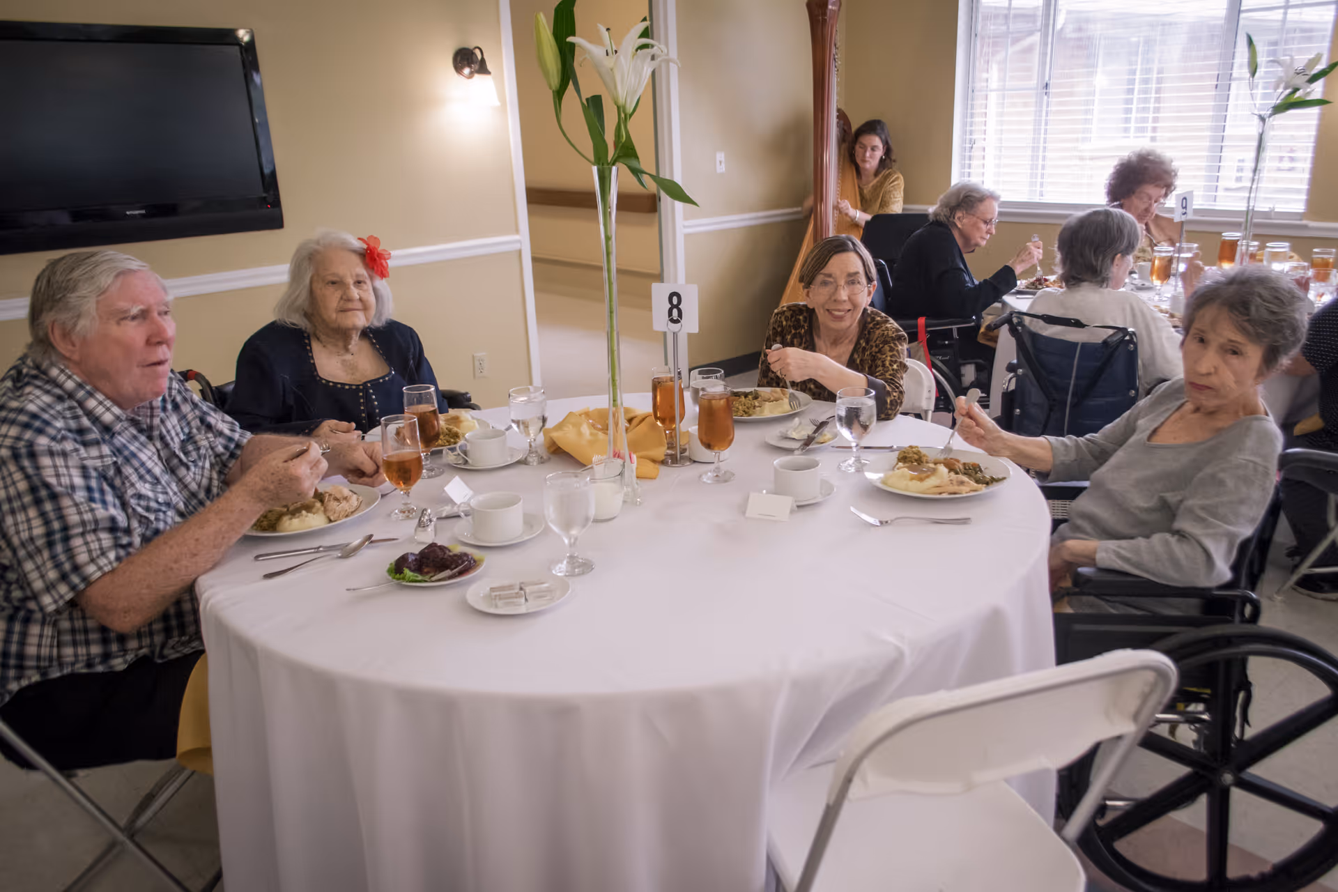 A group of elderly people sitting around a round table in a dining room, eating a meal. The table is covered with a white tablecloth and has glasses of iced tea, water, and plates of food. There is a tall vase with white flowers in the center of the table. The room has beige walls, a wall-mounted TV, and windows with blinds letting in natural light.