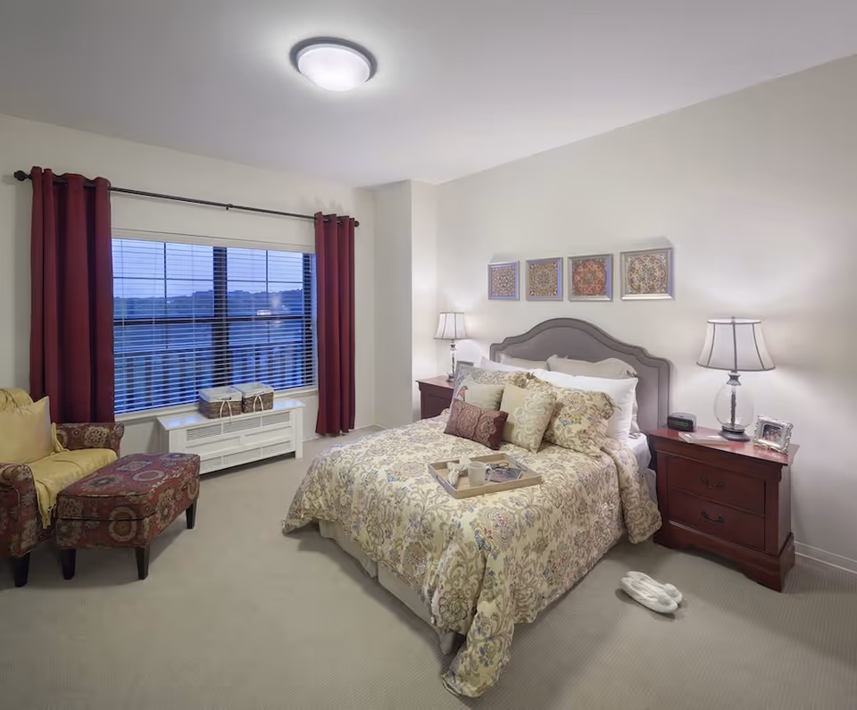 A cozy bedroom with a large bed covered in a floral patterned comforter and multiple pillows. There are two wooden nightstands on either side of the bed, each with a lamp. A pair of white slippers is placed on the carpeted floor next to the bed. A comfortable armchair with a matching ottoman sits near a large window with red curtains and blinds. The walls are light-colored with four framed decorative artworks above the bed.