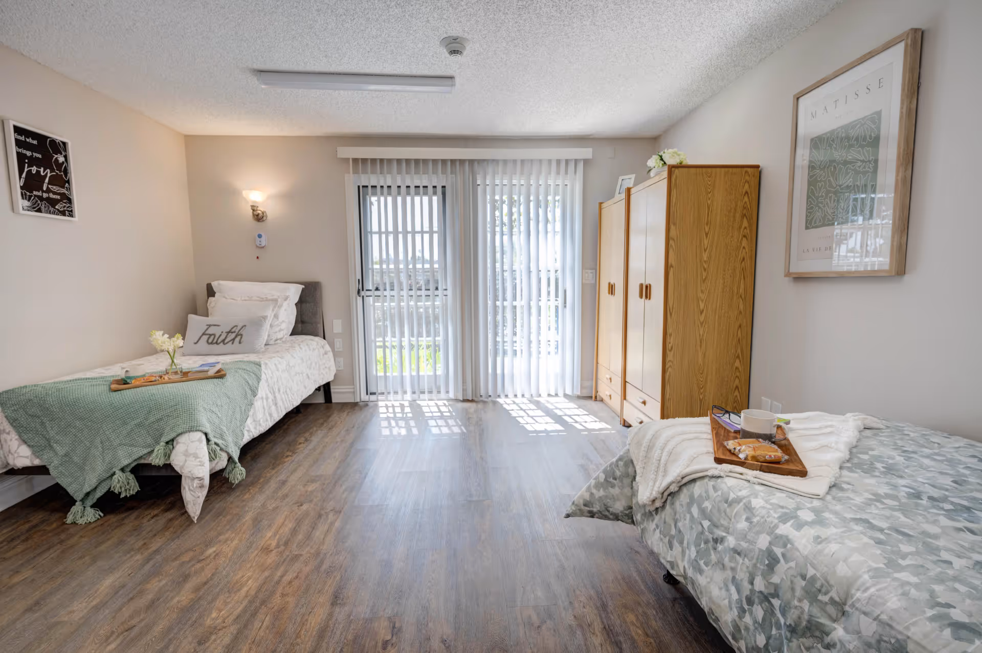 A bright and clean bedroom with two single beds on opposite sides of the room. Each bed is neatly made with decorative pillows and throws. A wooden wardrobe stands against the wall near a large window with vertical blinds letting in natural light. The room has wood-style flooring and light-colored walls adorned with framed artwork.