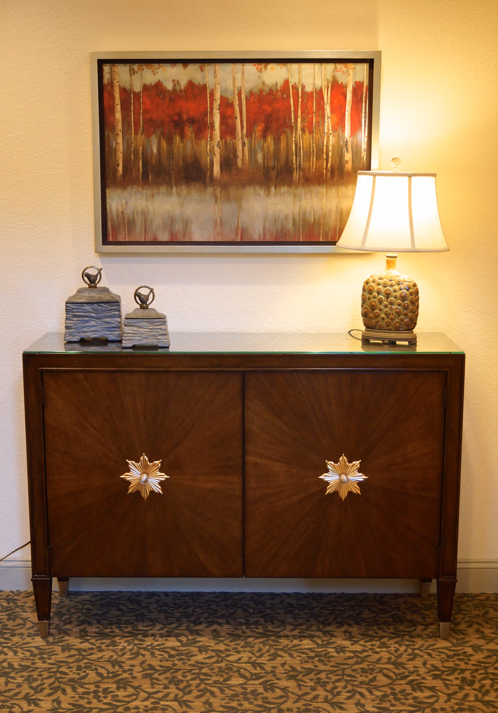 A wooden cabinet with decorative star-shaped handles, topped with two small ornamental boxes and a lamp with a patterned base and white lampshade. Above the cabinet hangs a framed painting of a forest with red and brown trees reflected in water.