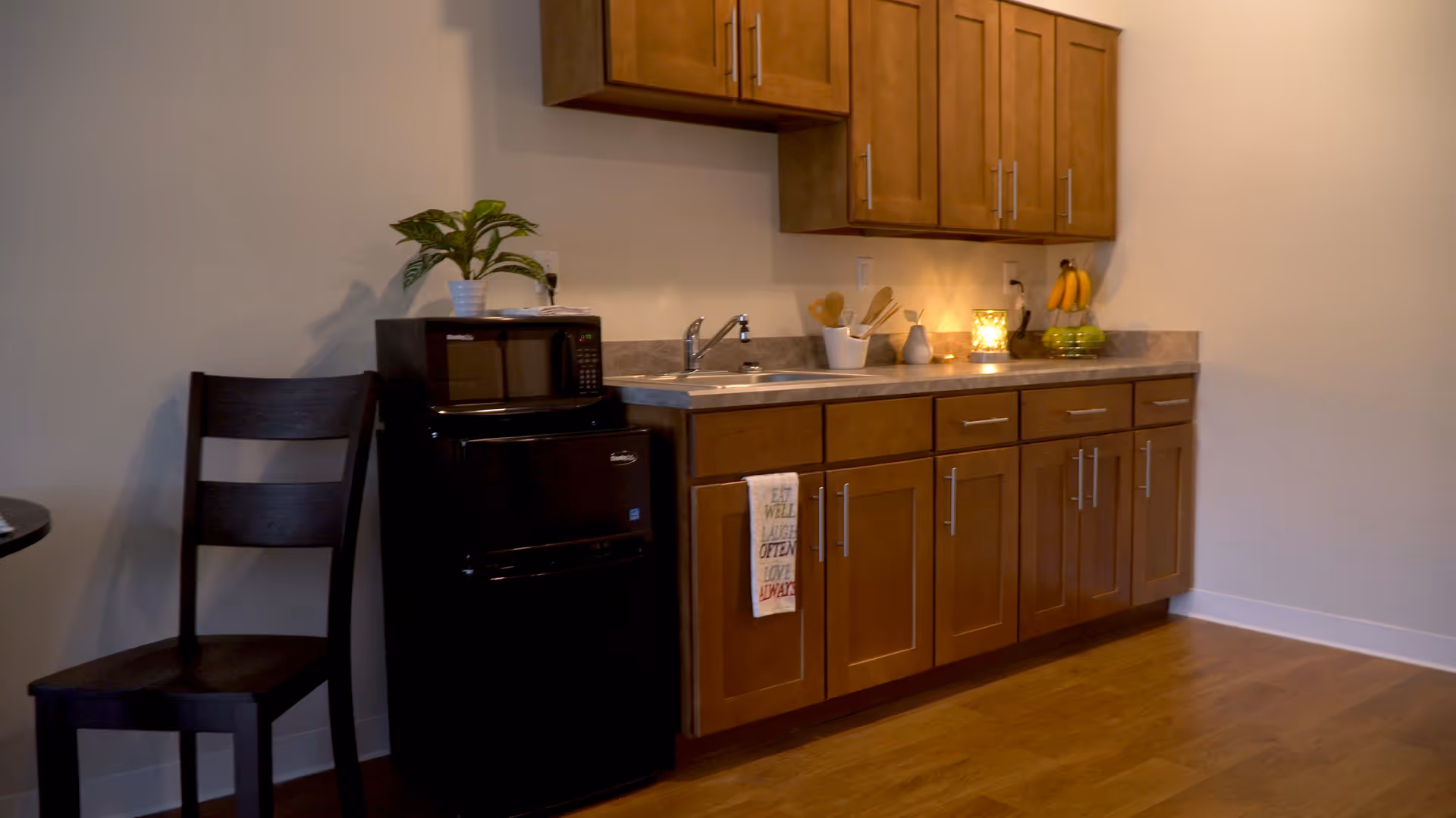 A small kitchen area with wooden cabinets, a countertop with a sink, a black microwave and mini fridge, a wooden chair, and some kitchen utensils and fruit on the counter.